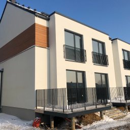 MAXI-BUD USŁUGI OGÓLNOBUDOWLANE MASZOTA HENRYK - Nowoczesny, dwukondygnacyjny dom z jasną elewacją, drewnianymi akcentami i czarnymi balustradami balkonowymi w zimowej scenerii, ukazujący efekt wykonanych prac.