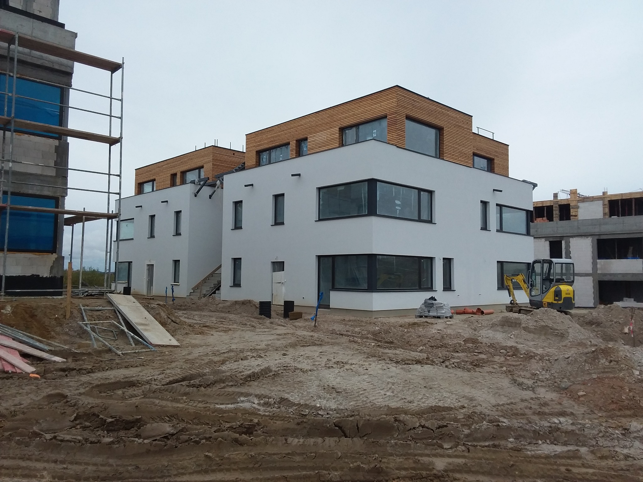 Nowoczesny, dwukondygnacyjny budynek z białymi ścianami i drewnianą elewacją na poddaszu, w trakcie budowy, z widocznym sprzętem budowlanym i rusztowaniami.