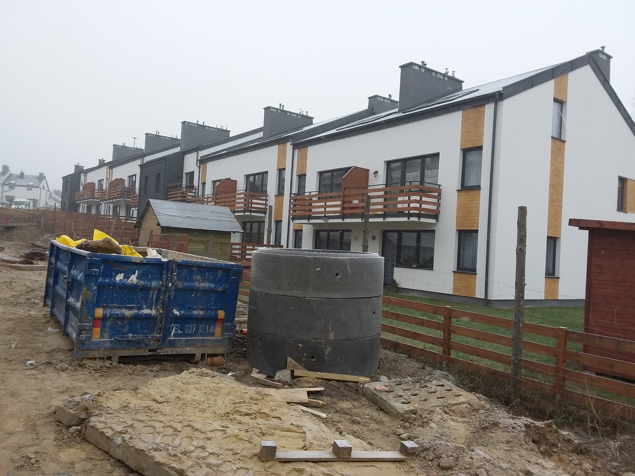 Nowoczesne domy szeregowe z balkonami, wykończone białą elewacją z drewnianymi akcentami. Na pierwszym planie kontener i betonowe kręgi na placu budowy.