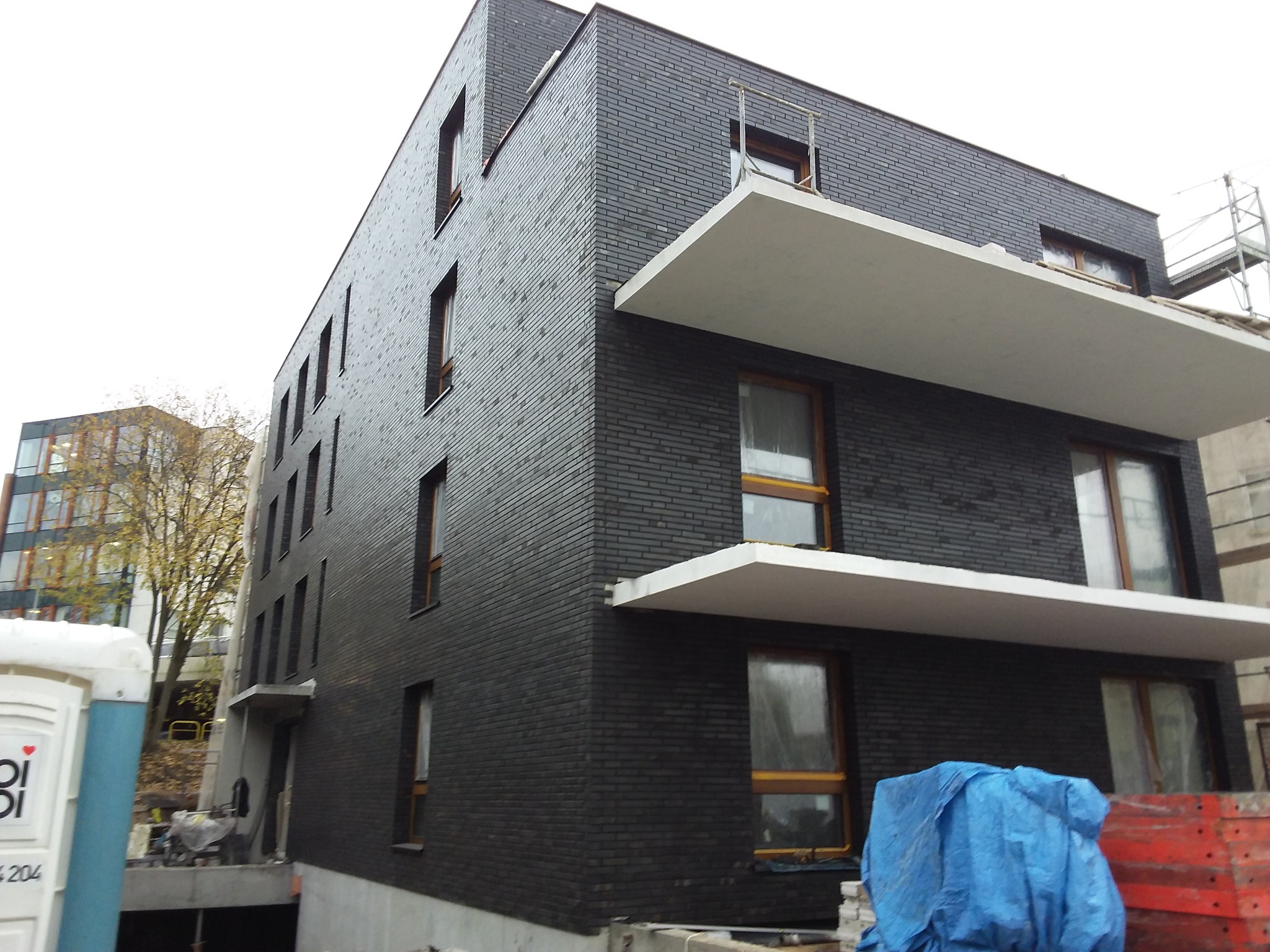 Nowoczesny budynek z ciemnej cegły z białymi balkonami, w trakcie budowy. Widoczne rusztowania i materiały budowlane. Ujęcie z dołu, podkreślające geometrię budynku.