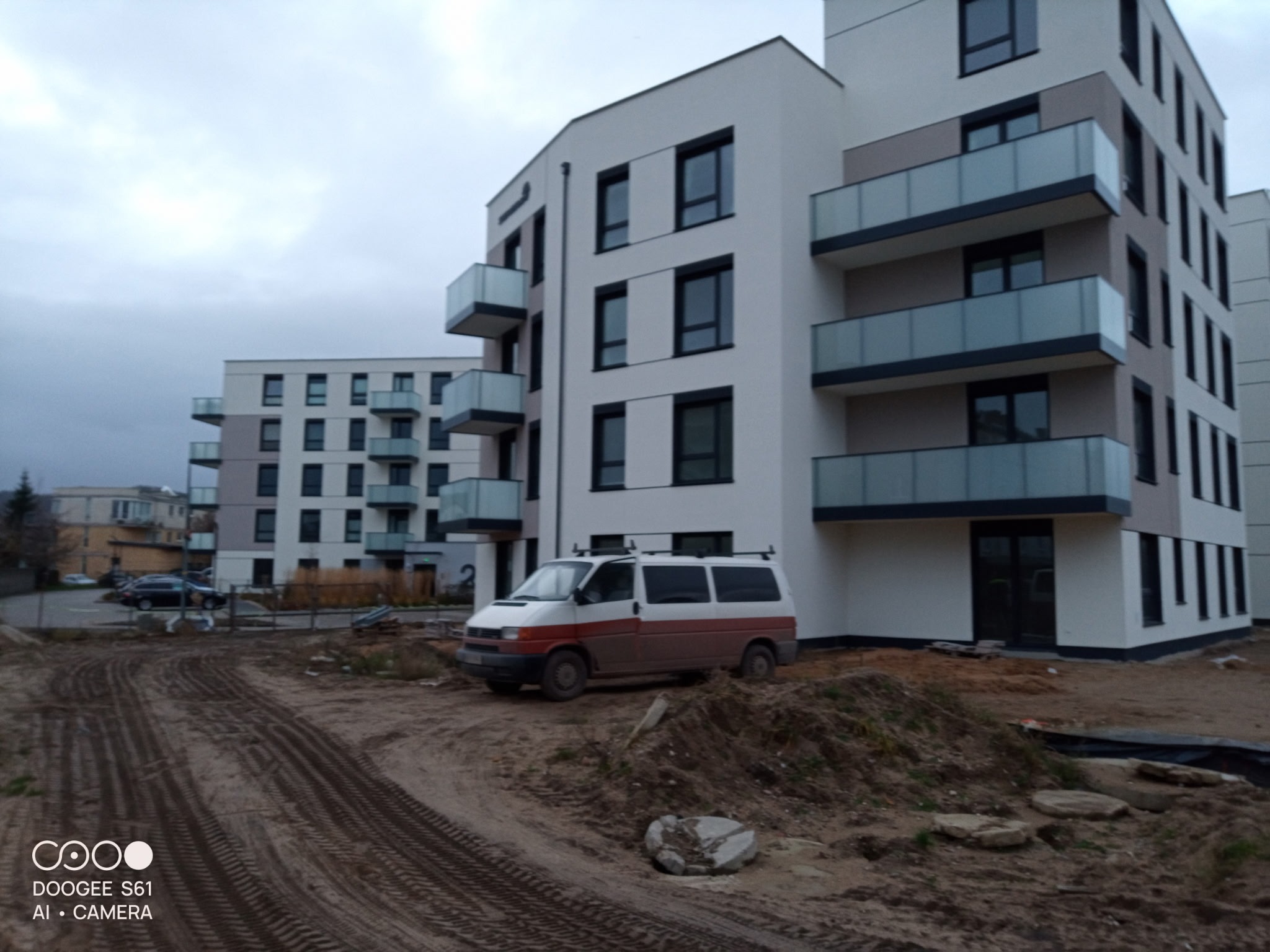 Nowoczesny budynek wielorodzinny z balkonami, w tle drugi budynek, na pierwszym planie bus na placu budowy. Szaro-biała elewacja, czarne okna.