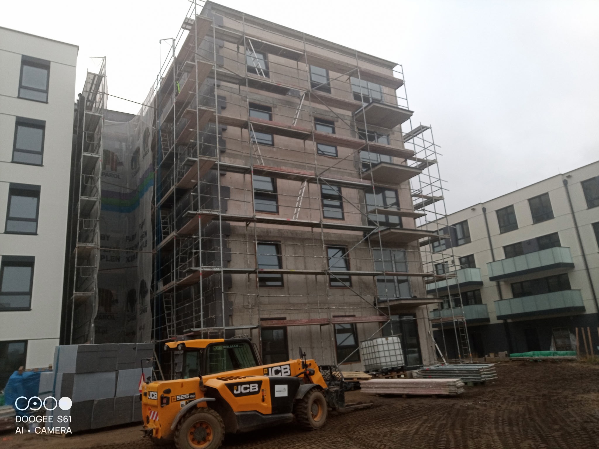 Wielopiętrowy budynek w trakcie ocieplania, otoczony rusztowaniami. Na pierwszym planie żółta ładowarka JCB i materiały budowlane. Widoczne są sąsiednie budynki.