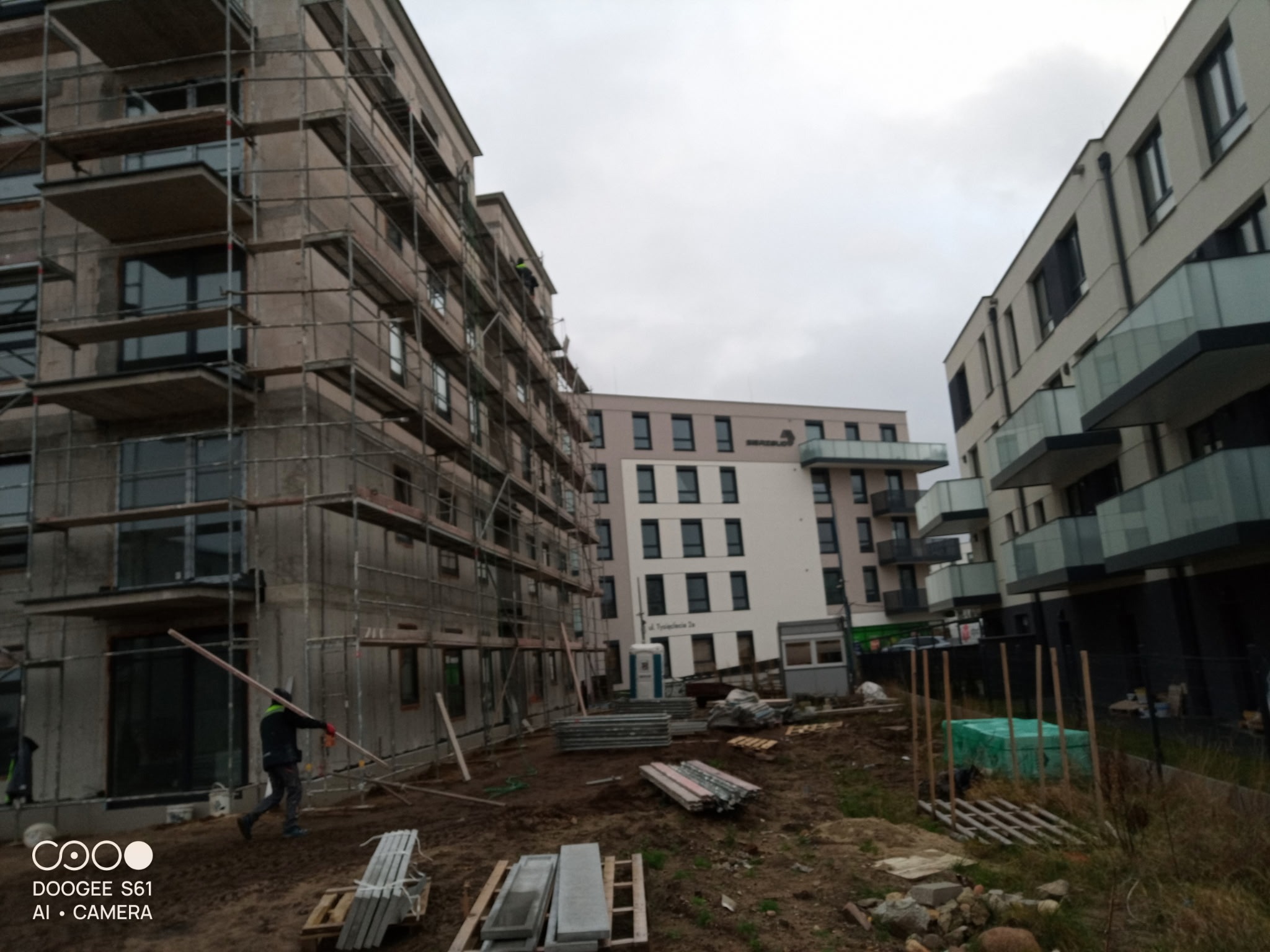 Budynek w trakcie prac elewacyjnych, widoczne rusztowania i materiały budowlane. Pracownik w trakcie pracy. Pochmurne niebo w tle.