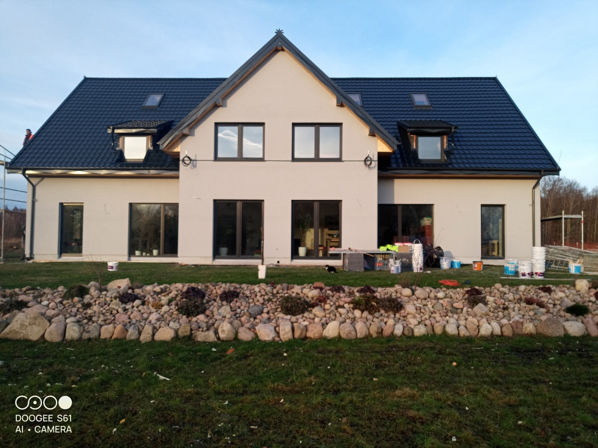 Nowoczesny dom jednorodzinny z ciemnym dachem i jasną elewacją, otoczony zielonym trawnikiem i kamiennym obrzeżem. Widoczne farby i rusztowanie sugerują prace wykończeniowe.