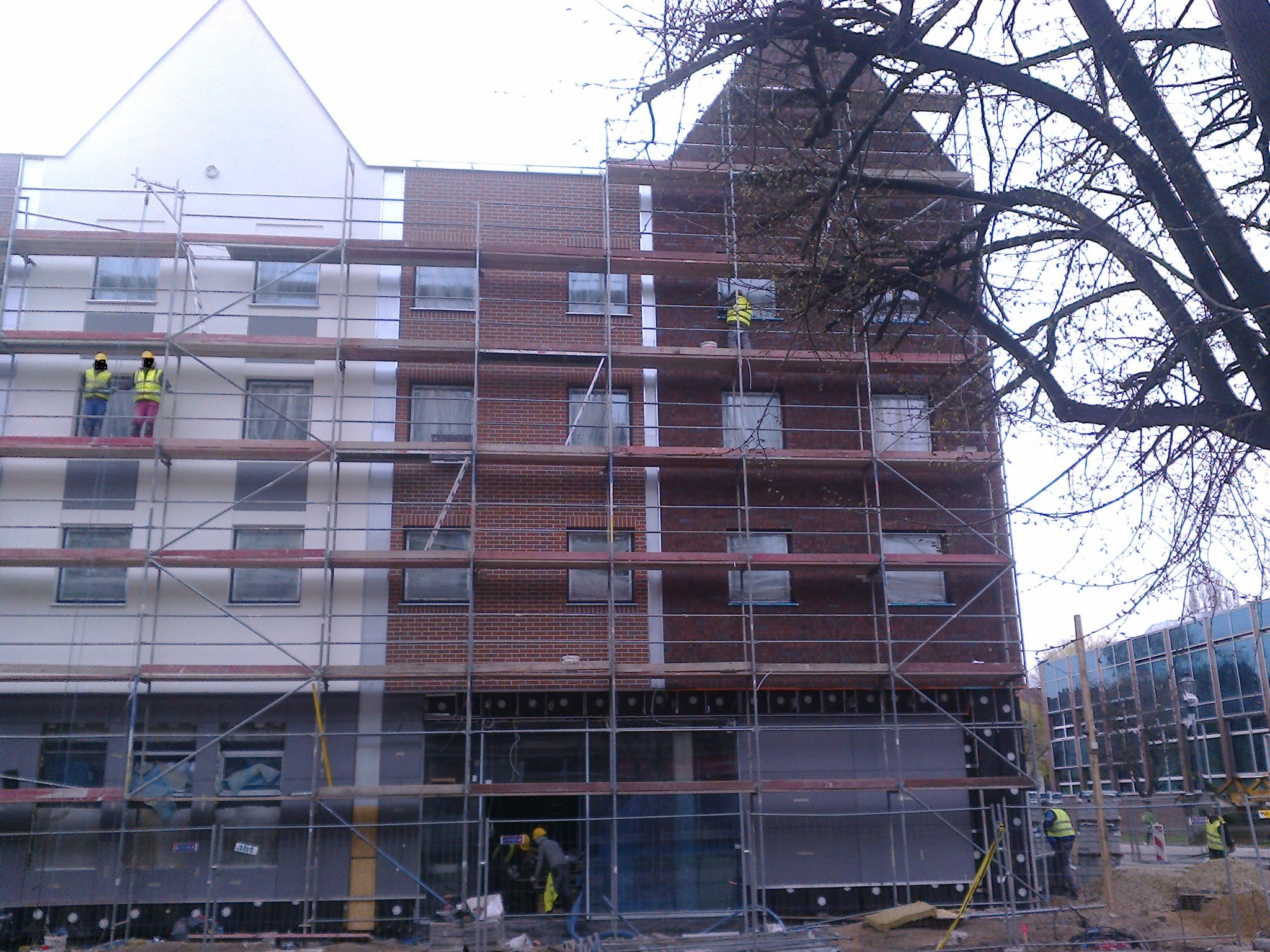 Budynek w trakcie prac elewacyjnych z rusztowaniem. Część fasady ocieplona, druga w surowej cegle. Pracownicy w żółtych kamizelkach na rusztowaniu.