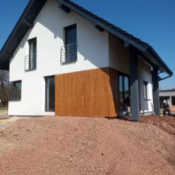 Nowoczesny dom jednorodzinny z białymi ścianami i elementami z drewnopodobnej okładziny elewacyjnej, widoczny stos czerwonego gruzu przed budynkiem i pracujący robotnicy w tle.