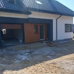 Nowoczesny dom jednorodzinny w trakcie budowy: czarny dach z dachówką, biała elewacja z ciemnoszarymi akcentami, drewniana brama garażowa częściowo otwarta, teren wokół domu przygotowywany...