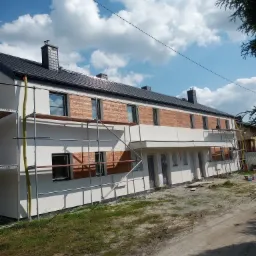 Dwukondygnacyjny budynek w Jastrzębiu-Zdroju w trakcie prac elewacyjnych, z widocznymi rusztowaniami i fragmentami ocieplenia imitującego drewno na elewacji.