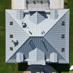 DACH-POL K&M OLKO SPÓŁKA JAWNA - Widok z góry na szary, metalowy dach z licznymi oknami dachowymi, kominami i wywietrznikami, otoczony zielonym trawnikiem, z logo firmy w prawym dolnym rogu.