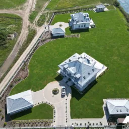 Widok z lotu ptaka na posesję z kilkoma budynkami z szarymi dachami z blachy trapezowej, otoczoną zielonym trawnikiem i ogrodzeniem, z samochodem na podjeździe i logo firmy w rogu.