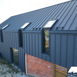 DACH-POL K&M OLKO SPÓŁKA JAWNA - Nowoczesny dom jednorodzinny z ciemnoszarą blachą na dachu i elewacji, widoczne okna dachowe i fragment muru z cegły klinkierowej w dolnej części elewacji.