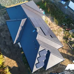 Widok z góry na nowy, ciemnoszary dach z dachówki ceramicznej z fragmentami paneli fotowoltaicznych. Widoczne krawędzie dachu, komin wentylacyjny i okno dachowe. Otoczenie to uprawne pole...