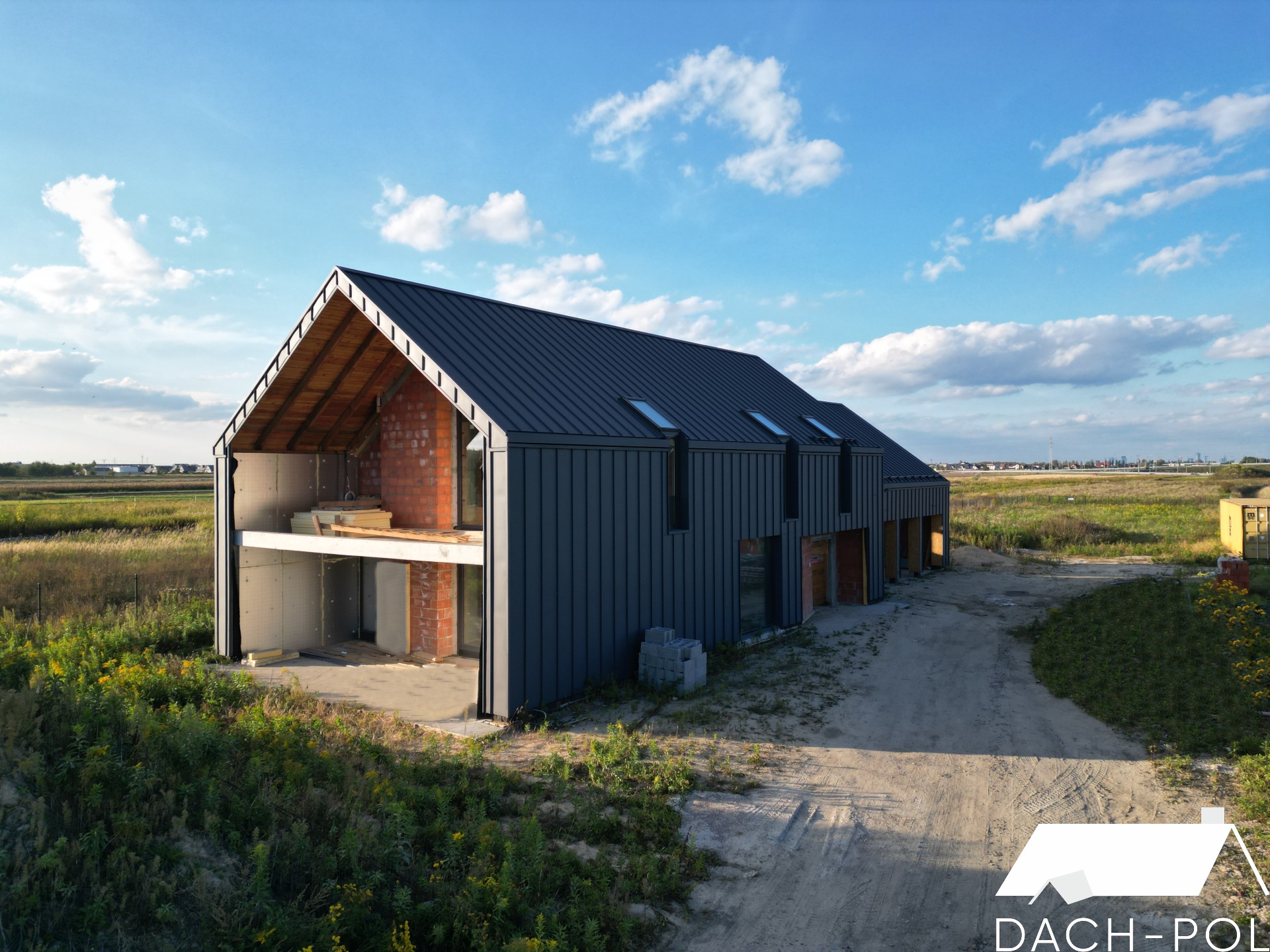 Nowoczesny dom w trakcie budowy z ciemną blachą na dachu i odsłoniętą konstrukcją więźby dachowej, widok z lotu ptaka na tle łąk i błękitnego nieba.