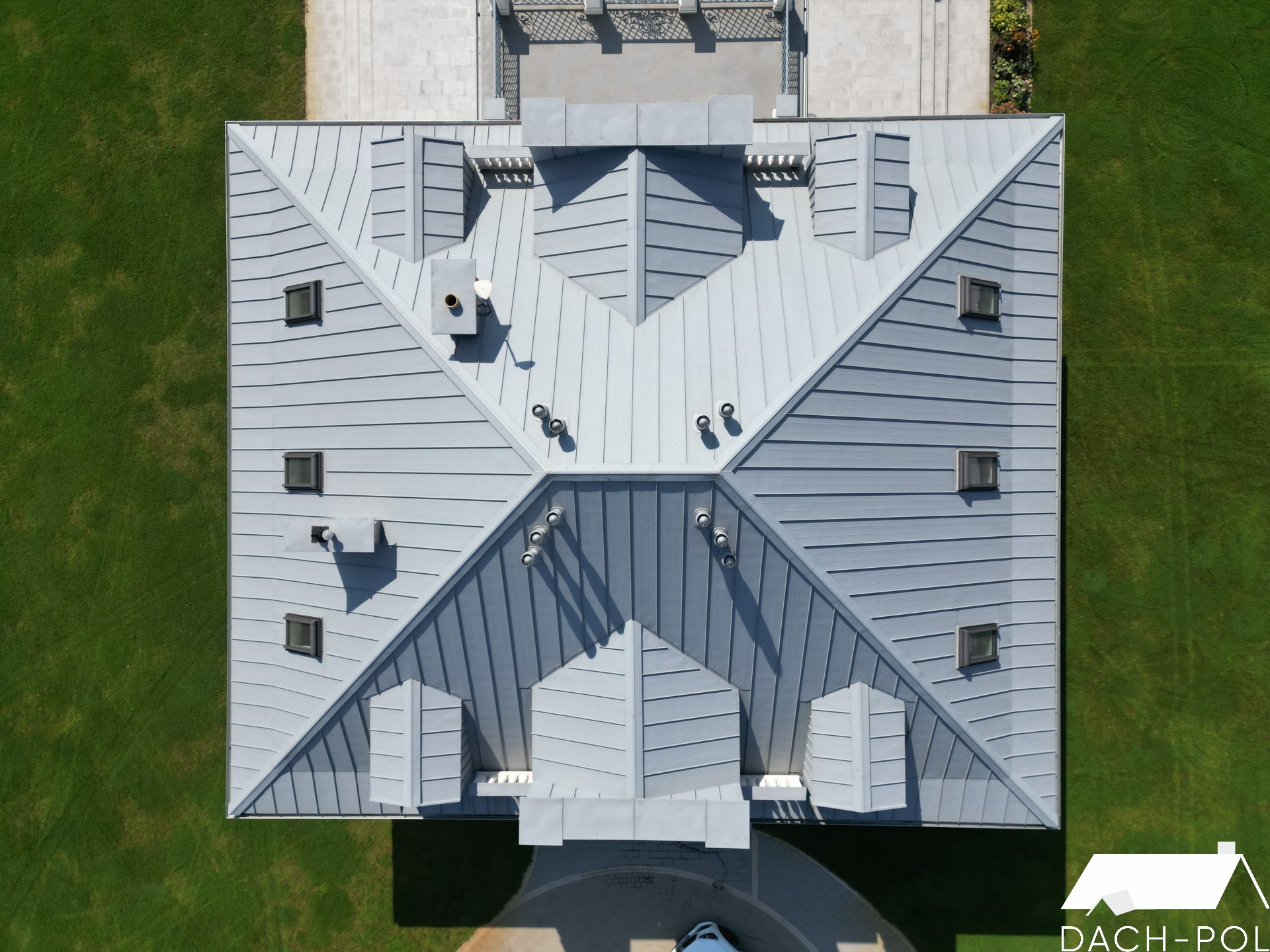 Widok z góry na szary, metalowy dach z licznymi oknami dachowymi, kominami i wywietrznikami, otoczony zielonym trawnikiem, z logo firmy w prawym dolnym rogu.