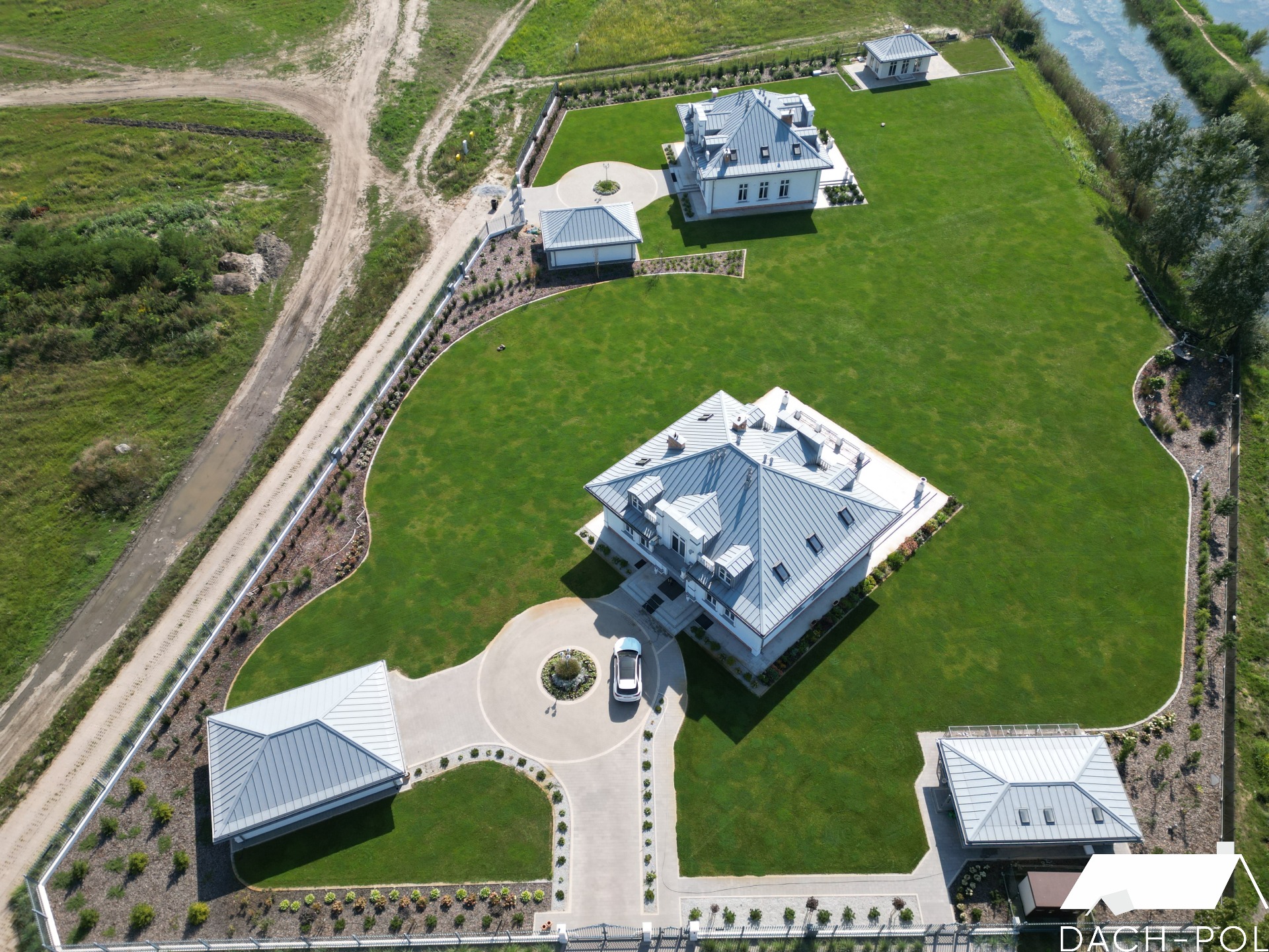 Widok z lotu ptaka na posesję z kilkoma budynkami z szarymi dachami z blachy trapezowej, otoczoną zielonym trawnikiem i ogrodzeniem, z samochodem na podjeździe i logo firmy w rogu.