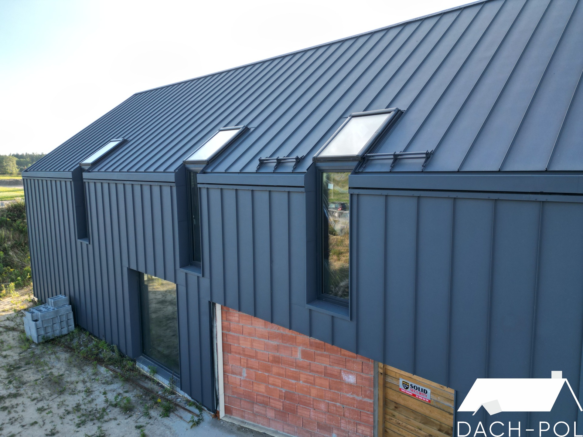 Nowoczesny dom jednorodzinny z ciemnoszarą blachą na dachu i elewacji, widoczne okna dachowe i fragment muru z cegły klinkierowej w dolnej części elewacji.