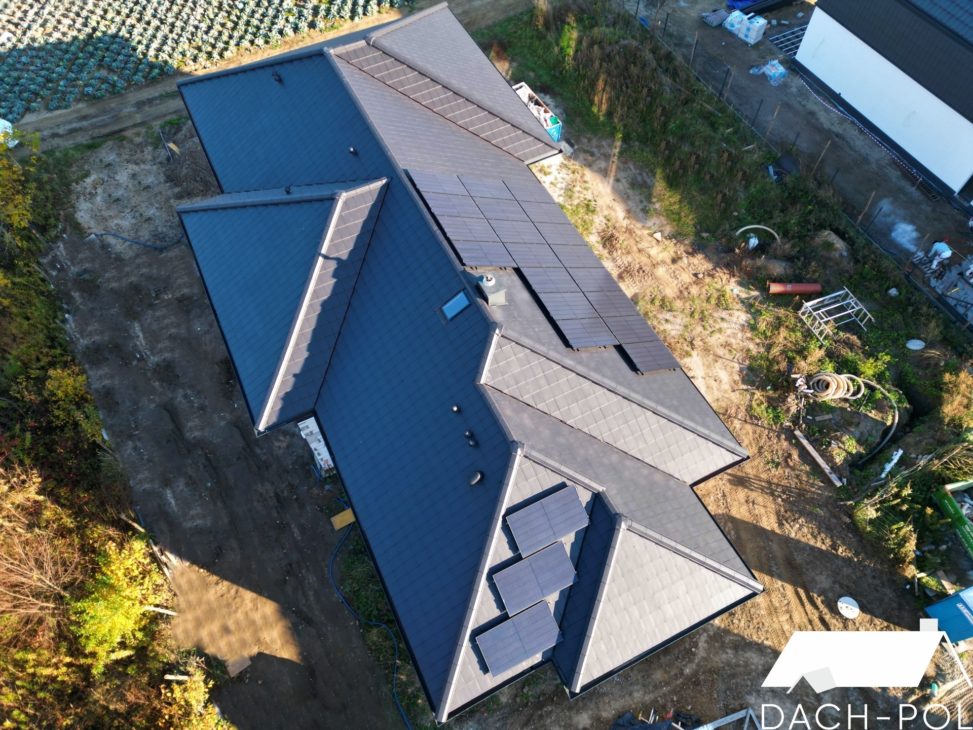 Widok z góry na nowy, ciemnoszary dach z dachówki ceramicznej z fragmentami paneli fotowoltaicznych. Widoczne krawędzie dachu, komin wentylacyjny i okno dachowe. Otoczenie to uprawne pole...