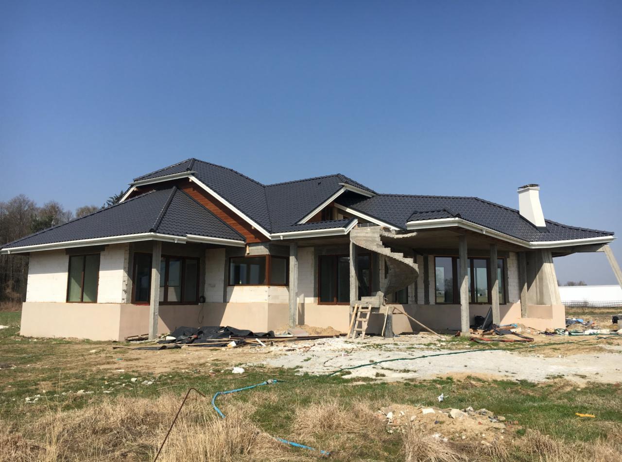 Dom jednorodzinny w trakcie budowy, z częściowo ukończonym dachem z ciemnej dachówki ceramicznej i spiralnymi schodami zewnętrznymi w surowym stanie, widoczne rusztowania i materiały budowlane.