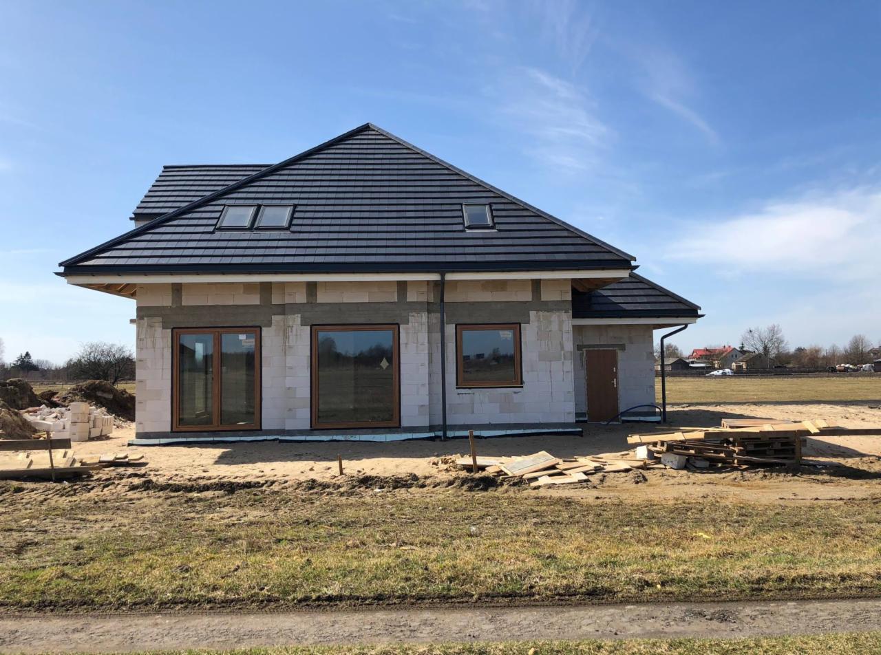 Dom w trakcie budowy z ciemną dachówką, widoczne okna z drewnianymi ramami, białe bloczki konstrukcyjne i materiały budowlane na ziemi.