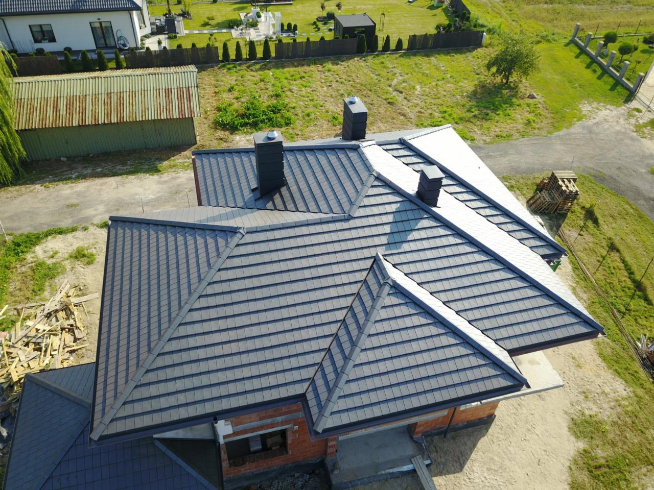Widok z lotu ptaka na dom z nowym, grafitowym dachem z dachówki ceramicznej, z trzema kominami, częściowo wykończony, z paletą dachówek obok i stertą drewna na podwórku.