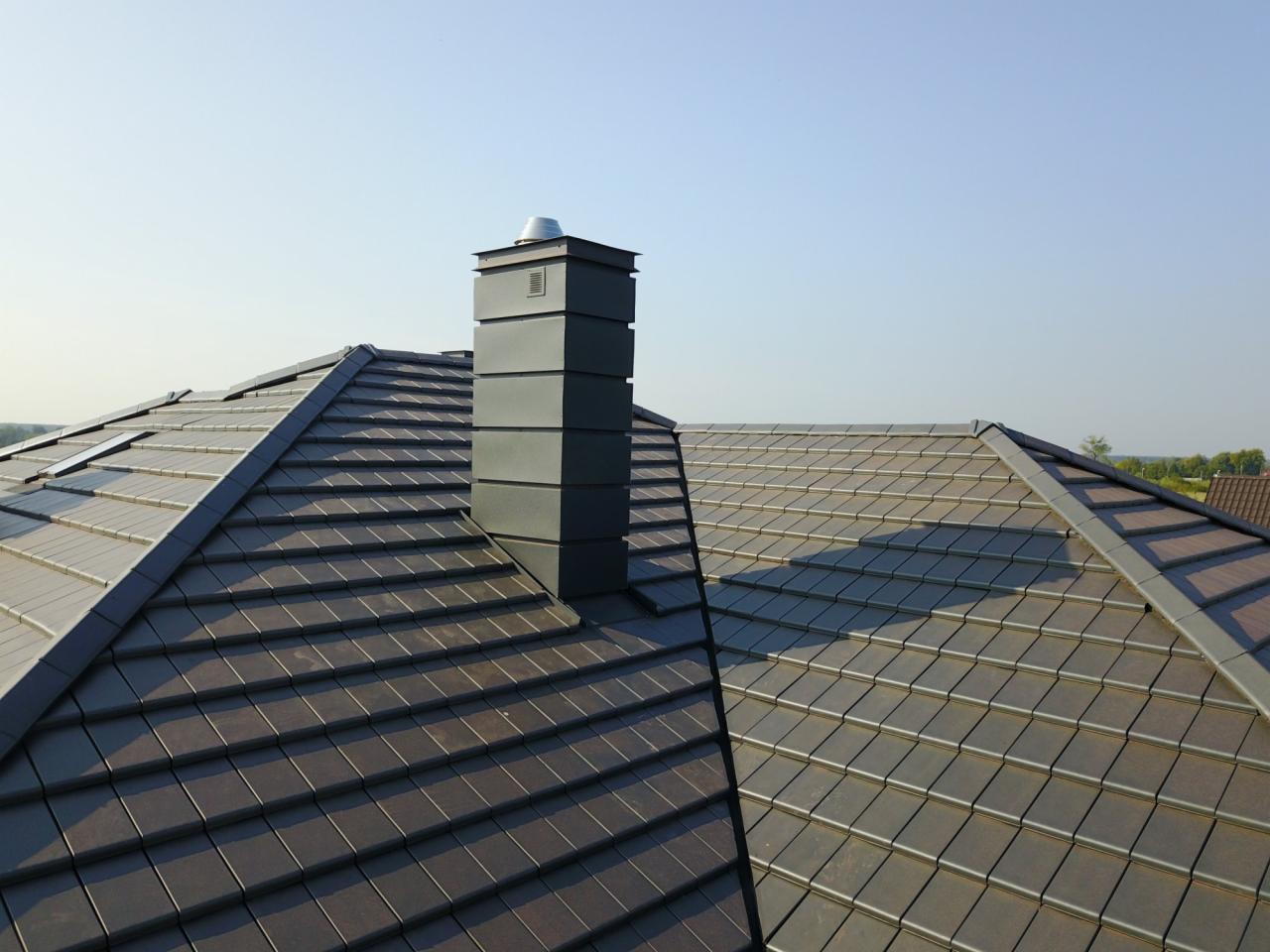 Nowoczesny dach z ciemnej dachówki ceramicznej i komin wentylacyjny z metalu, widok z lotu ptaka, błękitne niebo w tle.