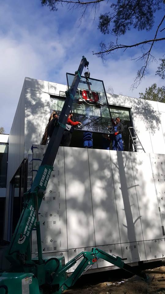 Montaż panelu elewacyjnego na budynku z wykorzystaniem dźwigu, widoczni pracownicy w strojach roboczych.