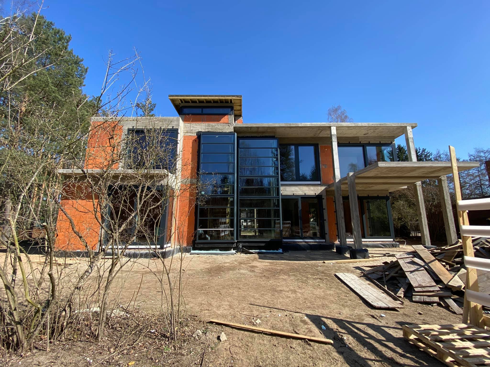 Surowy stan nowoczesnego domu z dużymi oknami i betonowym dachem w trakcie budowy, widoczne cegły i elementy konstrukcyjne, na pierwszym planie gałęzie drzew.