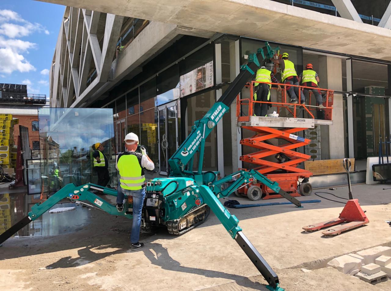 Montaż szklanych paneli fasadowych na budynku biurowym przy użyciu mini-żurawia gąsienicowego i podnośnika nożycowego, pracownicy w kamizelkach odblaskowych