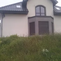 Elewacja domu jednorodzinnego po remoncie z widocznymi oknami z roletami zewnętrznymi i rusztowaniem z boku budynku, zarośnięta trawa na pierwszym planie.