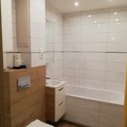 Nowoczesna łazienka z białymi płytkami imitującymi marmur, drewnopodobnymi akcentami na zabudowie toalety i szafce umywalkowej, oraz minimalistyczną wanną z frontem w pasującym stylu.