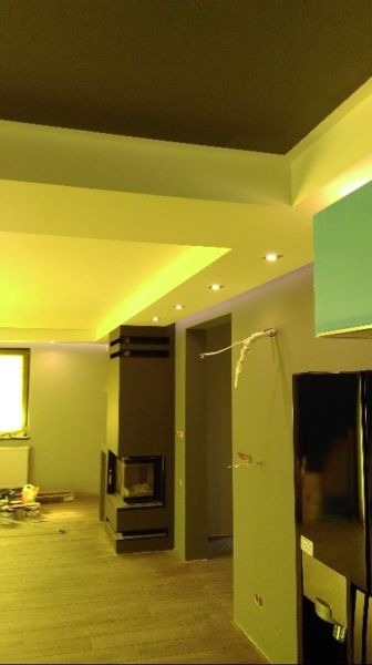 Wnętrze w trakcie prac wykończeniowych: pomalowane ściany na szaro, zamontowany kominek z czarnymi elementami, podwieszany sufit z oświetleniem punktowym, widoczne narzędzia i materiały budowlane...