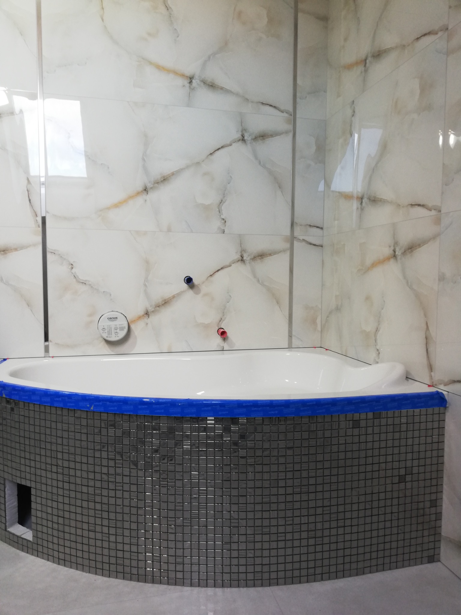 Wanna obudowana mozaiką w trakcie prac wykończeniowych, zabezpieczona niebieską taśmą malarską, z widoczną instalacją wodną na ścianie wyłożonej płytkami imitującymi marmur.