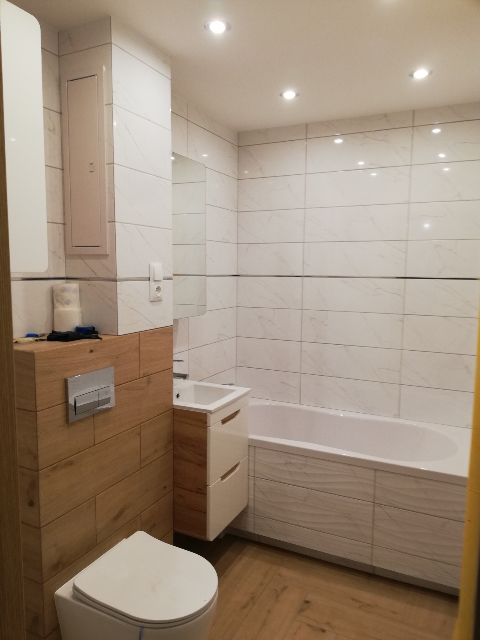 Nowoczesna łazienka z białymi płytkami imitującymi marmur, drewnopodobnymi akcentami na zabudowie toalety i szafce umywalkowej, oraz minimalistyczną wanną z frontem w pasującym stylu.