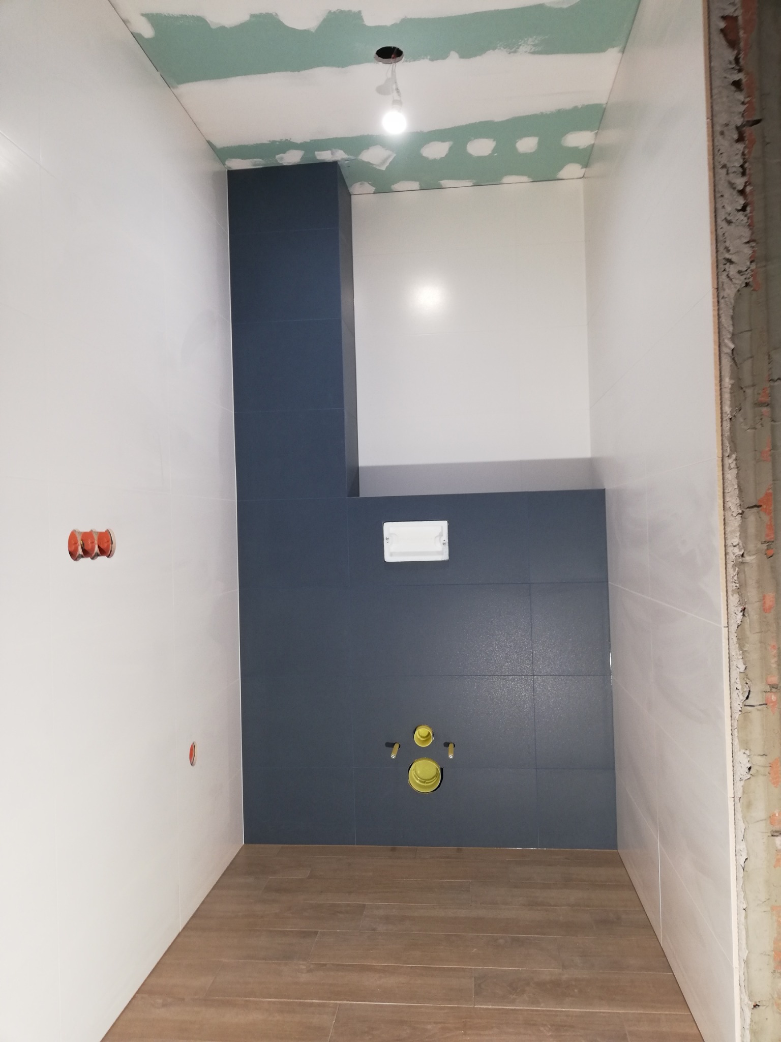 Wnętrze łazienki w trakcie remontu: częściowo wykończone ściany z białych i grafitowych płytek, drewnopodobna podłoga, sufit z płyt gipsowo-kartonowych przed malowaniem, widoczne instalacje...