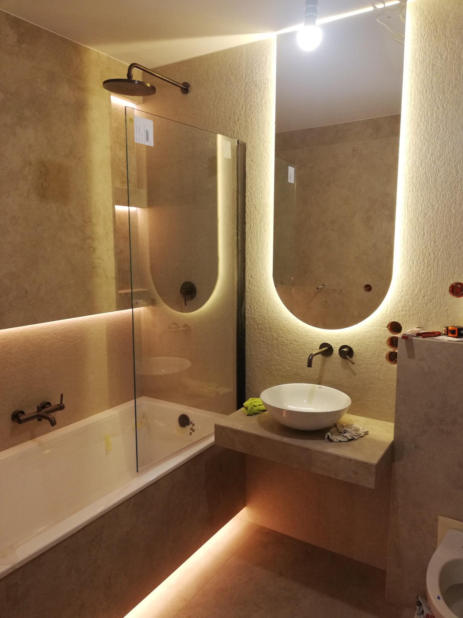 Nowoczesna łazienka z podświetlanym lustrem i kabiną prysznicową typu walk-in, wykończona beżowymi płytkami. Widoczna umywalka nablatowa i bateria w kolorze brązu.