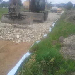 Caterpillar układa kamienisty materiał na geowłókninie, formując podłoże drogi; w tle widoczny walec drogowy i hałda ziemi.