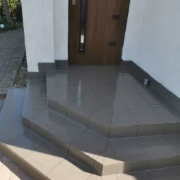 Wykończone płytkami schody wejściowe do domu, z ciemną drewnianą stolarką drzwiową i jasnym tynkiem elewacyjnym.