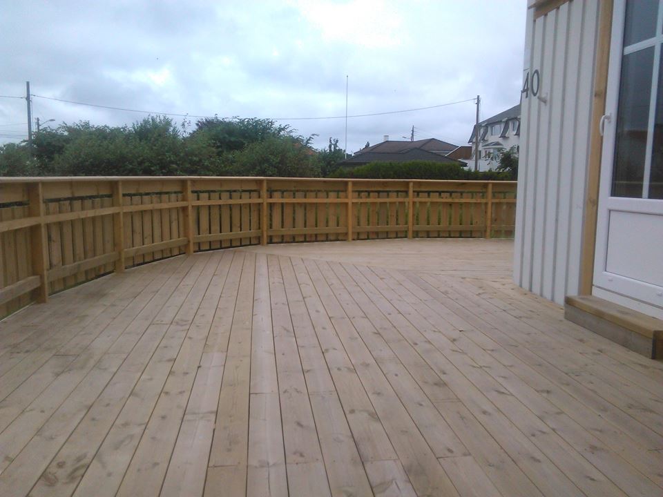 Drewniany taras z jasnego drewna otoczony płotem z pionowych desek, przylegający do białego budynku z numerem 40, widok na zieleń i domy w tle.