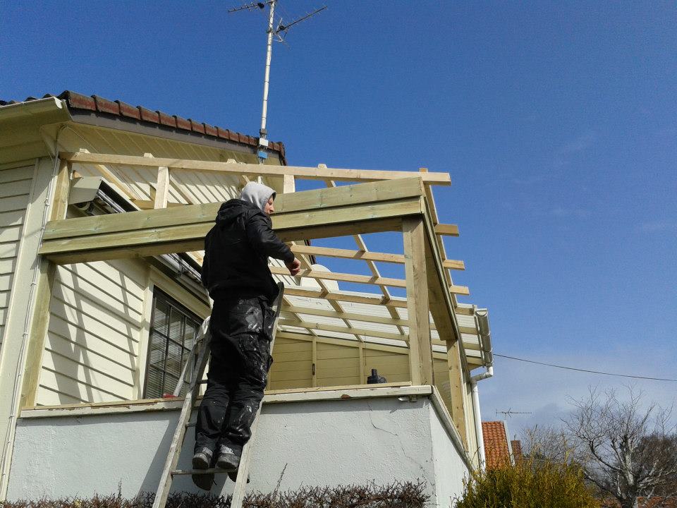 Pracownik na drabinie montuje drewnianą konstrukcję rozbudowy domu, widoczne elementy szkieletu i fragment elewacji budynku z jasną boazerią.
