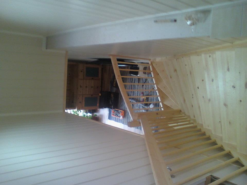 Wnętrze domu w trakcie rozbudowy, widoczne drewniane schody prowadzące na wyższą kondygnację, ściany wykończone boazerią, częściowo pomalowane na biało.