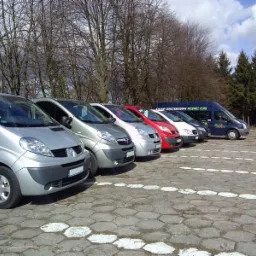 Rząd srebrnych, czerwonych i białych busów osobowych na parkingu z kostki, z napisem 'Krajowy i międzynarodowy przewóz osób' na jednym z pojazdów.