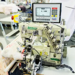 Profesjonalna maszyna szwalnicza Yamato z panelem sterowania, szyjąca materiał w kwiaty, w tle hala produkcyjna z pracownikami.