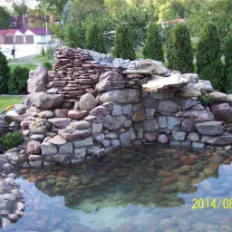 Kaskada wodna z kamieni w ogrodzie, z wyraźnym odbiciem w wodzie. W tle zielone tuje i budynki. Data na zdjęciu: 2014/08/27.