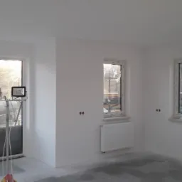 Wnętrze pokoju po szpachlowaniu ścian i sufitu, z widocznymi dwoma oknami, grzejnikiem i lampami halogenowymi na statywie.