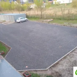 Wykończony podjazd z szarego kruszywa, obrzeża z jasnych betonowych elementów, dwa samochody zaparkowane na powierzchni, widok z góry.