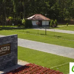 Widok na teren rekreacyjny z budynkiem i ścieżkami wyłożonymi szarą kostką, zielony trawnik, znak 'Rancho Stefanów' z kamienną podstawą i rabata z czerwonymi kwiatami na pierwszym planie.