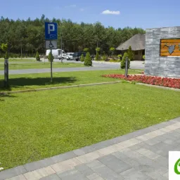 Parking ekologiczny z trawiastą nawierzchnią wzmocnioną kratką, obrzeża z szarej kostki, w tle znak parkingowy dla niepełnosprawnych i budynek z drewnianą strzechą.