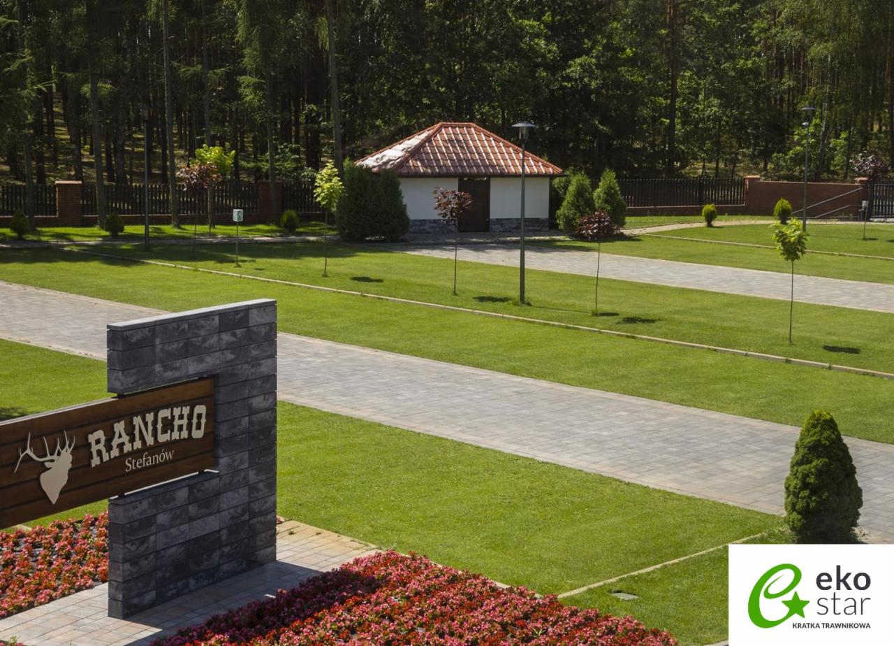 Widok na teren rekreacyjny z budynkiem i ścieżkami wyłożonymi szarą kostką, zielony trawnik, znak 'Rancho Stefanów' z kamienną podstawą i rabata z czerwonymi kwiatami na pierwszym planie.