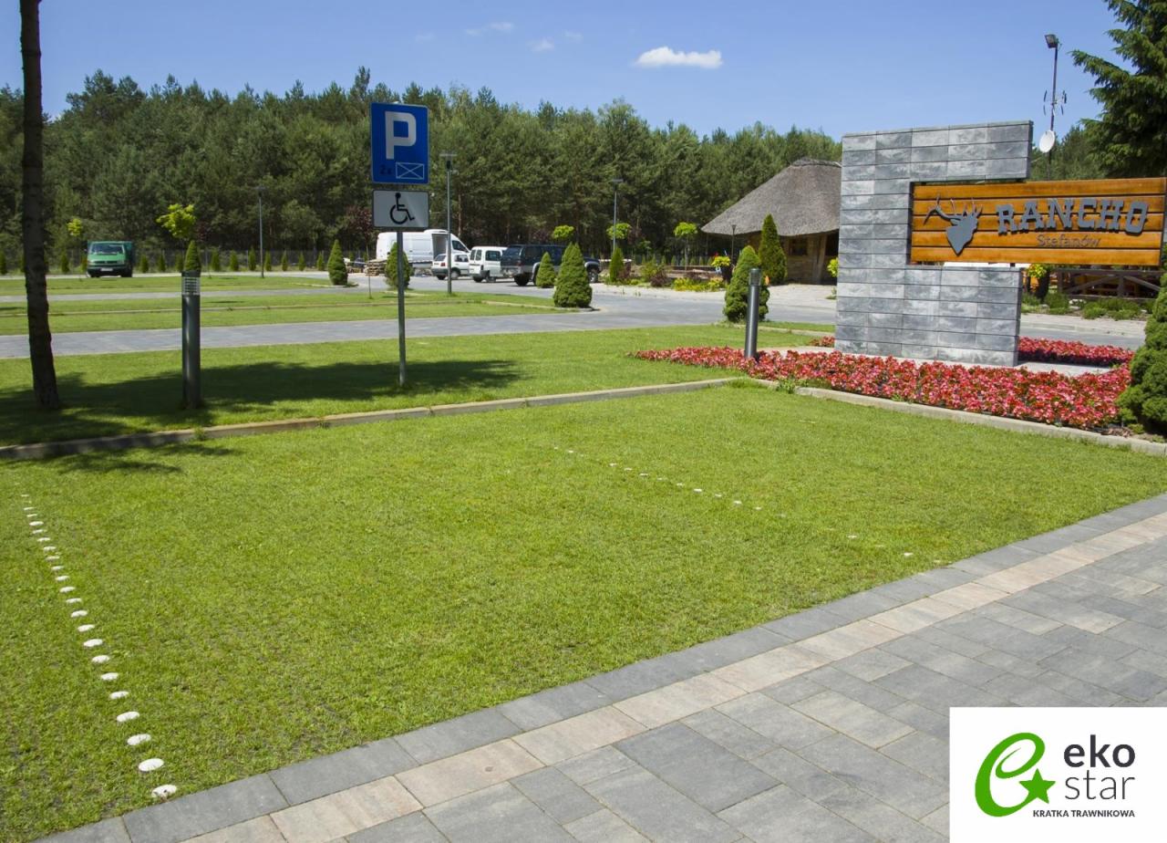 Parking ekologiczny z trawiastą nawierzchnią wzmocnioną kratką, obrzeża z szarej kostki, w tle znak parkingowy dla niepełnosprawnych i budynek z drewnianą strzechą.