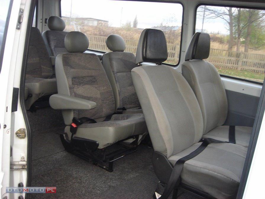 Wnętrze busa z szarymi fotelami pasażerskimi, widoczne pasy bezpieczeństwa i zagłówki, widok z otwartych drzwi na rzędy siedzeń.